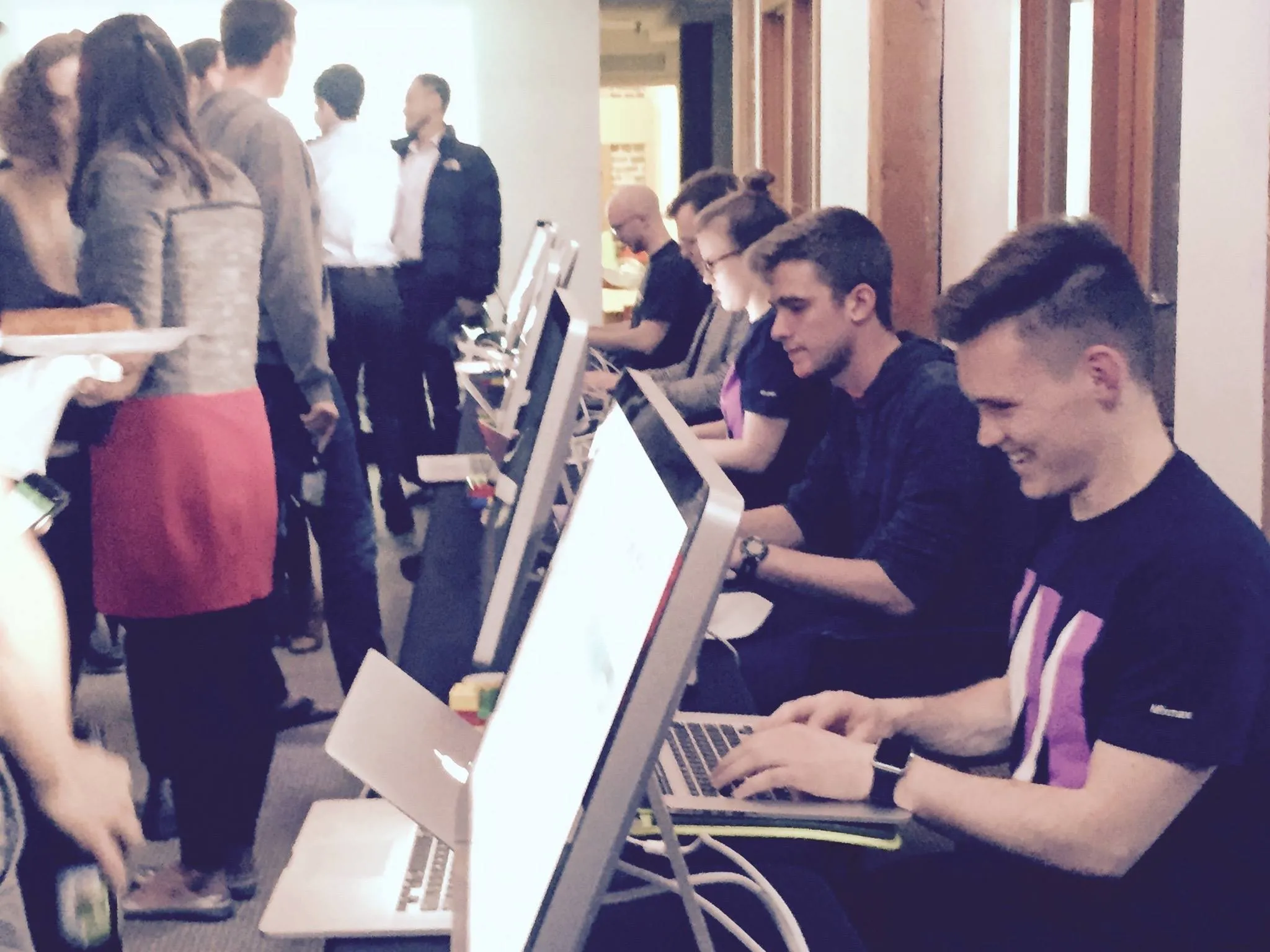 DareDev contestants coding side by side on laptops in a row while the audience mingles behind them
