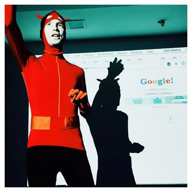 A host in a red morphsuit presenting a contestant's rebuild of the Google homepage projected on the wall
